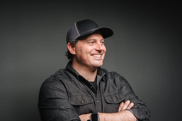 Side angle shot of Ryan Lucas with arms crossed and smiling. He's wearing a black baseball cap and a dark grey denim shirt.